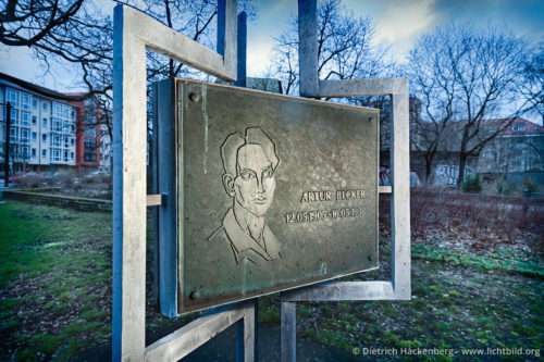 Artur Becker Gedenktafel in Hohenschönhausen - Im Ehrenhain zwischen Konrad-Wolf-/Obersee-/Degnerstraße, 13053 Berlin-Hohenschönhausen - Sicht auf eine von drei Tafeln in Obersee-/Konrad-Wolf-Straße in Berlin-Alt-Hohenschönhausen, dem Ortsteil in dem Artur Becker von 1929-1933 wohnte. Aufschrift Rückseite: „im Zuchthaus Burgos (Spanien) von den Faschisten ermordet“. Foto © Dietrich Hackenberg Der Kommunist Artur Becker war der jüngste Reichstagsabgeordnete der Weimarer Republik. Er starb als Interbrigadist in Burgos, vermutlich an den Verletzungen, die er sich während eines Gefechtes zugezogen hat. In der DDR wird er zum Helden für die Jugend stilisiert und man dichtet ihm einen Heldentod auf dem Hof eines Foltergefängnisses der Gestapo im spanischen Burgos an. […] Er dachte an die Passionaria an ihr Wort: ‚Lieber im Stehen sterben als auf den Knien leben!’ Ein stolzes ein spanisches Wort. […] Als sie von ihm forttraten und das erste Kommandowort erscholl, fetzte er die Binde herunter und rief: Seht her – so stirbt ein...’ […] Mit Recht trägt die höchste Auszeichnung, die unsere Freie Deutsche Jugend für besondere Leistungen verleiht, die Bezeichnung ‚Artur Becker Medaille’. […] Der Mann fiel als seine Idee siegte. Wir wissen um unseren Sieg. Es ist gut von den Menschen zu wissen, die ihn vorbereiten halfen. […] Aus den letzten Seiten und dem Nachwort eines propagandistischen DDR-Jugendbuches über Artur Becker. Greulich. ...und nicht auf den Knien. Roman von dem streitbaren Leben des Artur Becker