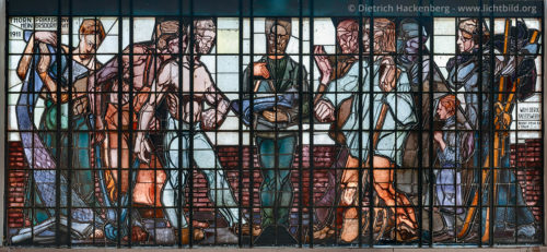 Bahnhofsfenster von Jan Thorn Prikker - Hagen Hauptbahnhof - Auf Betreiben des Kunstsammlers Karl Ernst Osthaus wurde der neue Hagener Hauptbahnhof 1911 mit einem monumentalen Glasgemälde ausgestattet. Osthaus hatte zielstrebig keinen heimischen Maler, sondern den niederländischen Künstler Johan Thorn Prikker (1868-1932) beauftragt. Das Glasfenster ist knapp 9 Meter breit und 3,60 Meter hoch. Metallbänder und Eisen teilen die Fläche in über 150 eckige Felder ein. Ausgeführt wurde es von einer Berliner Firma. Beim Verlassen des Hauptbahnhofs Richtung City, kann man das von Tageslicht bestrahlten Glasfenster gut erkennen. Im Zentrum der Malerei steht der Architekt als Lehrer und Anführer der Künstler mit Buch und Zirkel. Rechts und links stehen vor einer niedrigen Ziegelmauer Vertreter von Handel und Gewerbe: ein Maurer, ein Eisenhüttenmann, eine Weberin, ein Bauer und ein Grubenarbeiter. Dahinter sind noch drei weibliche Figuren unterschiedlichen Alters, deren Sinnbild nicht eindeutig ist. Foto © Dietrich Hackenberg