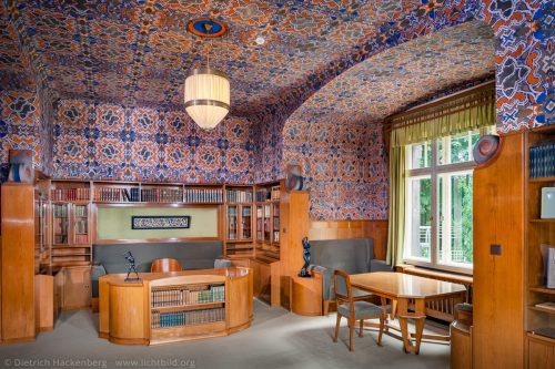 Arbeitszimmer von Karl Ernst Osthaus im Hohenhof. Das Arbeitszimmer ist Höhepunkt der Raumabfolge im Erdgeschoss. Die eingebauten Möbel aus grau polierter Esche hatten einen grünlichen Grundton. Gegen den Protest des Architekten van de Velde, der für die Raumdecke ein gebrochenes Weiß vorsah, ließ Osthaus 1911 eine in orange, graugrün und blau gehaltene Schablonenmalerei von Johan Thorn Prikker auftragen. Hagen