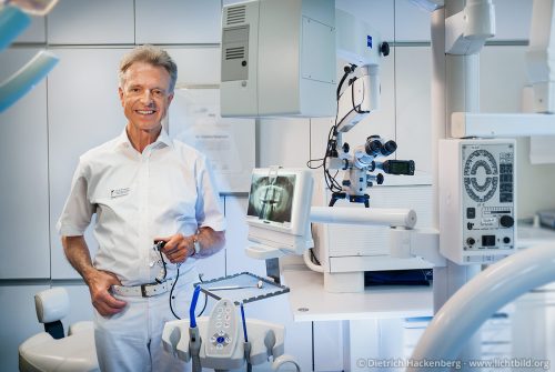 Ein Zahnarzt mit einer Lupenbrille in der Hand steht inmitten seiner Geräte. Der Zahnarzt wird als Behandler nach modernen Methoden dargestellt.
