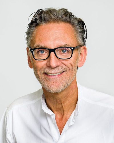 Der in Weiß gekleidete Zahnarzt ist als Porträt vor einem neutralen Hintergrund abgebildet.